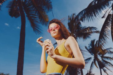 Sarı elbiseli şık bir kadın elinde dondurma tutuyor. Palmiyelerle çevrili, parlak mavi gökyüzüne karşı.