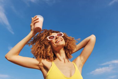 Sarı mayo giymiş neşeli bir kadın açık mavi gökyüzüne karşı smoothie ile kutlama yapıyor. Mutluluk ve yaz titreşimleri yayıyor.
