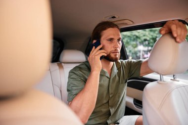 Uzun saçlı ve sakallı bir adam, yumuşak, nötr renklerle çevrili bir aracın arka koltuğunda arama yapıyor.