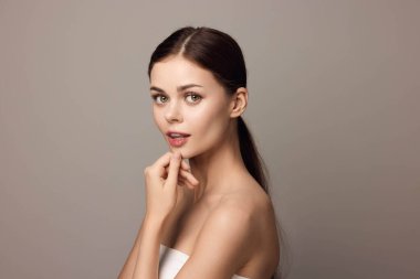 Çekici genç bir kadın, elleri çenesinde poz veriyor ve gri arka plandaki kameraya bakıyor. Kişisel markalaşma veya yaşam tarzı konsepti için kadınsı ve kendinden emin imaj.