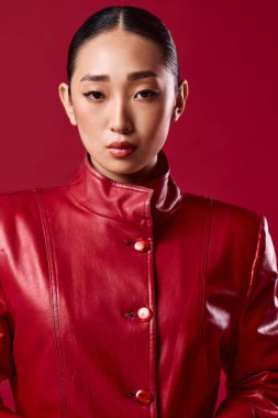 Kırmızı deri ceketli Asyalı esmer. Gözleri kırmızı arka planda, elleri kalçasında poz veriyor..