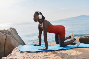 Kırmızı elbiseli ve kulaklıklı formda bir kadın, suyun kenarındaki kayalarla çevrili mavi bir mindere yoga pozu veriyor.
