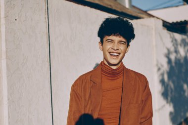 Pas rengi balıkçı yaka ve kahverengi ceket giyen neşeli genç bir erkek figürü, açık renkli bir duvar arka planına karşı neşeli bir şekilde gülümsüyor.