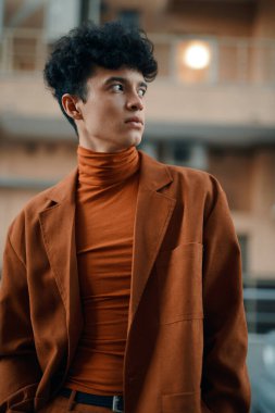 Zarif kahverengi ceketli genç bir adam ve balıkçı yakalı kazak yukarı bakıyor, yumuşak odaklı şehir arka planına karşı.