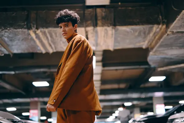 stock image A fashion forward male model showcases a brown outfit, exuding confidence in a dimly lit parking area, highlighting urban style and modern aesthetics