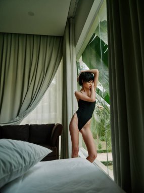Siyah mayo giymiş bir kadın modern bir yatak odasının geniş penceresinin yanında perdelerden süzülen yumuşak ışıkla düşünceli bir şekilde duruyor. Derin bir düşüncelere dalmış gibi görünüyor..