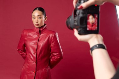 Kırmızı deri ceketli Asyalı esmer kadın aynı ceketi giyen başka bir Asyalı esmer kadının kırmızı arka planda fotoğrafını çekiyor..