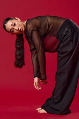 Siyah pantolonlu Asyalı bir kadın ve üstü açık bir şekilde kırmızı arka planda dizlerinin üstüne çöküp ayaklarını havaya kaldırıyor. moda konsepti.