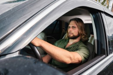 Arabadaki kaslı bir adam, araba sürerken kararlı görünüyor, penceresinin dışında palmiye ağaçları görünüyor, macera duygusu yayıyor.