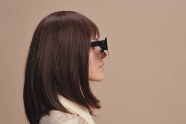 Uzun kahverengi saçlı, siyah güneş gözlüklü, beyaz gömlekli ve siyah ceketli bir kadın yan tarafa bakıyor.