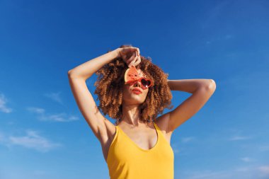 Kıvırcık saçlı kaygısız bir kadın yaz havasının tadını çıkarıyor, güneş gözlüğü ve bir dilim karpuz ile poz veriyor, neşe ve sıcaklığı yansıtıyor.