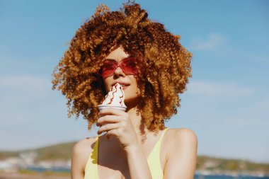 Şehvetli kıvırcık saçlı neşeli bir kadın dondurma külahını tutuyor, şık bir güneş gözlüğü takıyor, açık hava arka planına karşı koyuyor.