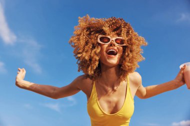 Kıvırcık saçlı ve güneş gözlüklü neşeli genç bir kadın kumsalda kutlama yapıyor, açık mavi gökyüzünün altında içki tutuyor.