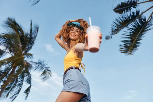 stock image A joyful woman in a yellow top and shorts smiles while holding a refreshing drink outdoors