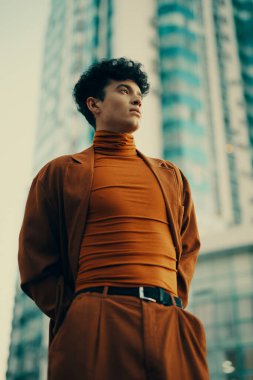 Kendine güvenen kahverengi elbiseli genç bir adam, çağdaş moda tarzını sergileyen, yüksek binaların arka planına karşı poz veriyor.