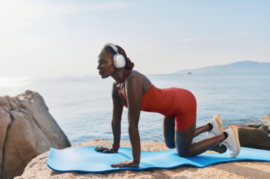 Güneşli bir günde dışarıda yoga yapan sağlıklı bir kadın, deniz manzarasının keyfini çıkarırken kulaklık takıyor sakin sular ve kayalık arka plan atmosferi güçlendiriyor.