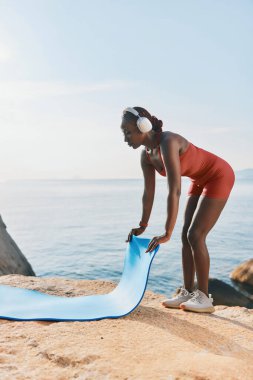 Formda bir kadın figürü kayalık bir yüzeyde mavi bir yoga minderi açıyor, sakin bir okyanus arkaplanı ve yumuşak güneş ışığıyla.