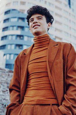 Kahverengi takım elbise ve balıkçı yaka giyen modacı erkek model çağdaş mimarinin arka planına gülümsüyor.