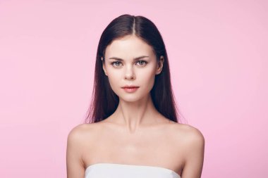 Pembe arka planda beyaz havluyla poz veren uzun kahverengi saçlı güzel bir genç kadın. Zarif ve kendinden emin görünüyor. Büyüleyici bir görüntü yaratıyor..