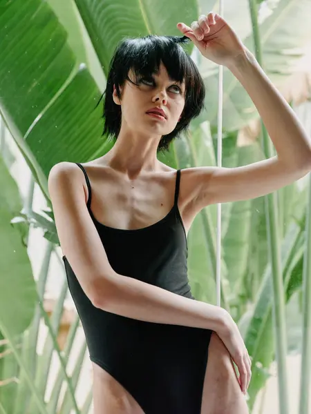 Stock image A contemplative woman in a black swimsuit stands against a lush, green tropical leaf background, exuding a sense of introspection and natural beauty.