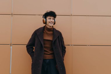 Kahverengi ceketli ve siyah balıkçı yakalı genç turuncu duvarın önünde duruyor, kulaklık takıyor. şık bir adamın moda portresi.