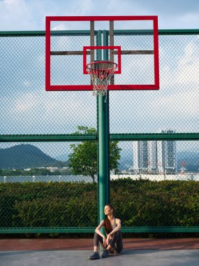 Spor giyimli bir kız, açık bir gökyüzüne ve şehir manzarasına karşı bir basketbol potası ile rahatlıyor.