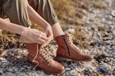 Kahverengi çizmeli, ayakları havada ve elleri bağcıklı bir kadın, ileri moda gezgini rahat ayakkabı güzelliğini keşfediyor