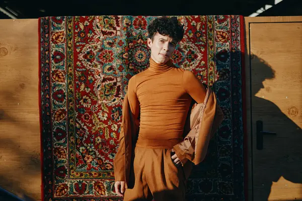 stock image A young male model dressed in a contemporary brown ensemble stands confidently in front of an ornate, colorful tapestry