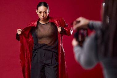 Kırmızı ceketli ve siyah pantolonlu Asyalı bir kadın kamerayla kırmızı arka planda fotoğraf çekiyor. moda konsepti.
