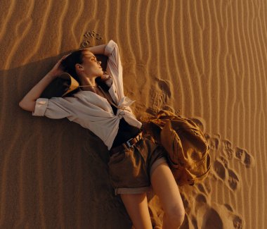 Çölde kumulda dinlenen yalnız bir kadın, açık mavi gökyüzünün altında kameraya geri dönüyor.