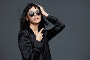 Siyah takım elbiseli, güneş gözlüklü, siyah elbiseli ve güneş gözlüklü bir kadın eli başının üstünde poz veriyor, yan yatırıyor, kendinden emin ve şık bir aura yakalıyor.