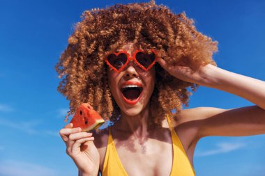 Kıvırcık saçlı, kalp şeklinde güneş gözlüğü takan neşeli bir kadın elinde parlak mavi bir gökyüzüne karşı bir dilim karpuz tutuyor.
