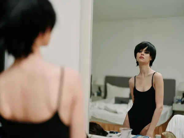 stock image Woman in black bodysuit looking thoughtfully at herself in the mirror, in a cluttered bedroom, reflecting on self-image and personal thoughts.