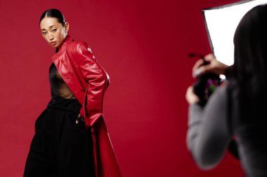 Kırmızı deri ceketli ve siyah pantolonlu moda Asyalı kadın elinde kamerayla kırmızı arka planda poz veriyor. Şık bir kıyafetle kendi fotoğrafını çekiyor..