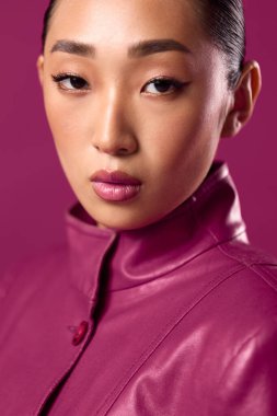 Mor deri ceketli, saçları at kuyruklu, pembe arka planda poz veren Asyalı bir kadın. Şık görünümlü şık bir esmer..