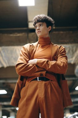 Çapraz kollu genç bir adam, aynı kahverengi elbiseyi giymiş, kırsal bir arka planda duruyor.