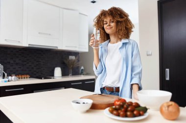 Mutfakta elinde bir bardak su ve bir tabak yemek tutan bir kadın.