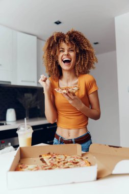 Kıvırcık saçlı genç bir kadın bir pizza kutusunun önünde pizzanın tadını çıkarıyor.