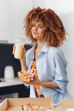 Kıvırcık saçlı, mutlu genç bir kadın bir dilim pizza ve sıradan bir ortamda içkinin tadını çıkarıyor.