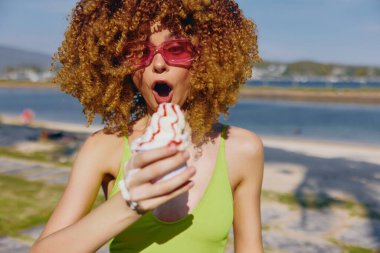 Neşeli bir kadın dondurma tutuyor, pembe güneş gözlüğü takıyor, kumdan ve denizden oluşan renkli bir geçmişi var.