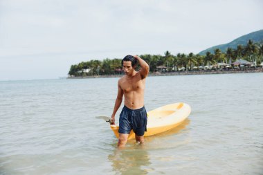 Serene Kayak Deneyimi: Turkuvaz sularıyla tropik bir sahilde yaz tatilinin tadını çıkaran mutlu bir Asyalı adam, Palm Trees 'in ortasında kanosuyla kürek çekiyor..