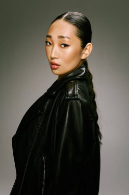 Asyalı kadın siyah deri ceket kamera grisi arka plan pozu veriyor. Modaya uygun giyinmiş genç bir kadının şık ve modaya uygun fotoğrafı..