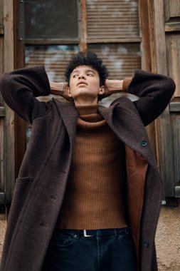 Kahverengi ceketli ve balıkçı yaka kazaklı genç adam elleri başının üzerinde pencereye yaslanmış kameraya bakıyor. şık ve şık bir fotoğraf..