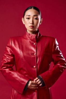 Kırmızı deri ceketli Asyalı esmer, elleri kalçasında ve ceplerinde kırmızı arka planda poz veriyor..