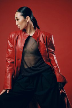 Kırmızı deri ceketli, siyah pantolonlu, kırmızı arka planda poz veren Asyalı kadın. Gözler kapalı, eller kalçada. Şık ve şık görünüm..
