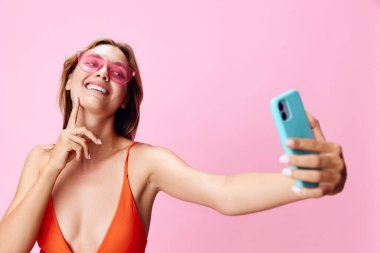 Canlı pembe mayo giymiş şık bir kadın renkli bir arka planda akıllı telefonuyla güneşli bir selfie çekiyor.