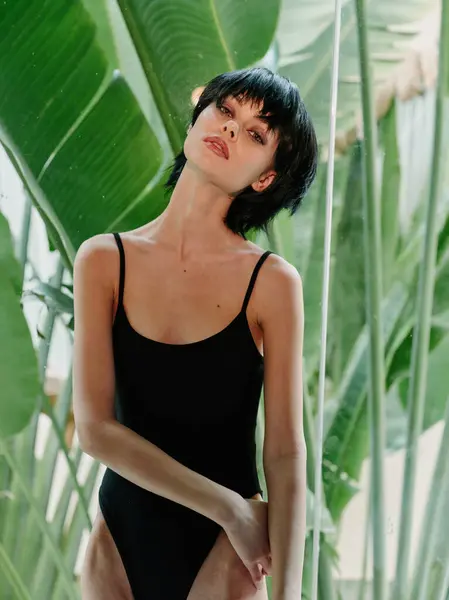 stock image Confident woman in a black bodysuit posing against lush tropical leaves, showcasing elegance and poise, with a serene expression and stylish short hair.