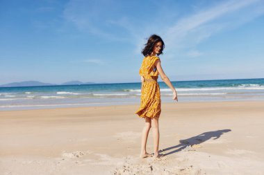 Süzülen sarı elbiseli güzel bir kadın zarif bir şekilde kumlu sahilde duruyor sakin okyanusla birlikte fon olarak.