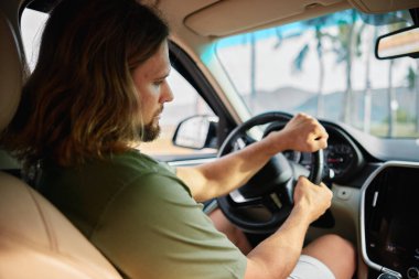 Sürücü koltuğundaki bir adamın yakın çekimi, direksiyonu kavraması, palmiye ağaçları ve arka planda parlak bir gökyüzü, macera hissi uyandırıyor.