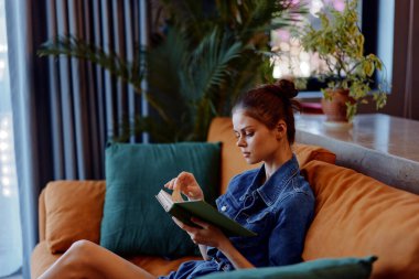 Sıcak bir oturma odasında, turuncu bir koltukta oturmuş kitap okuyan bir kadın.